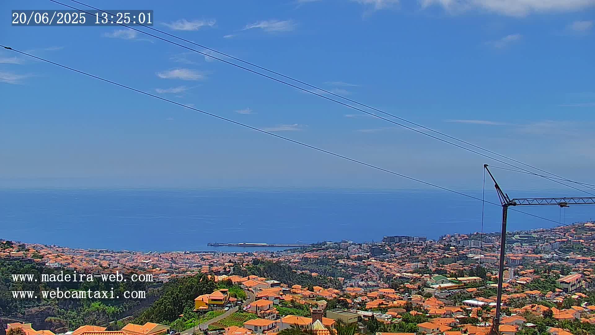 A coastal city with orange-roofed houses extends to a calm blue ocean under a mostly clear, sunny sky.