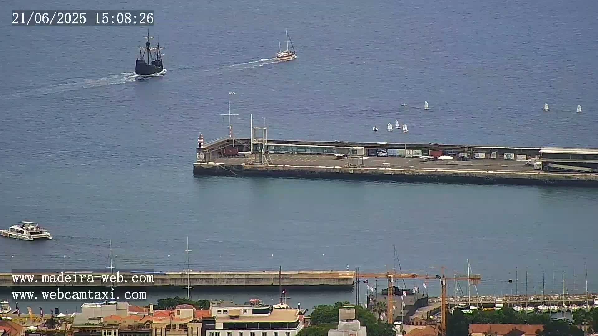 A large caravel-style ship and several sailboats are visible on a calm ocean near a pier and harbor with numerous docked boats and buildings in the distance.