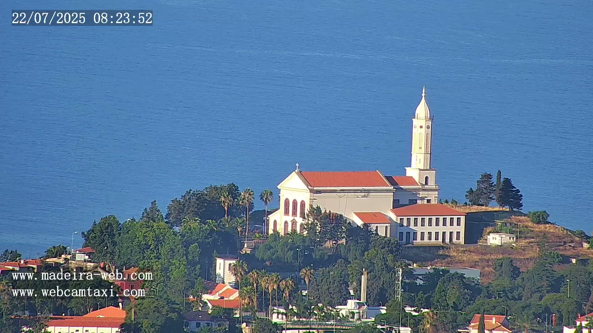 A white church with a tall tower sits atop a hill overlooking a calm blue ocean on a clear day.