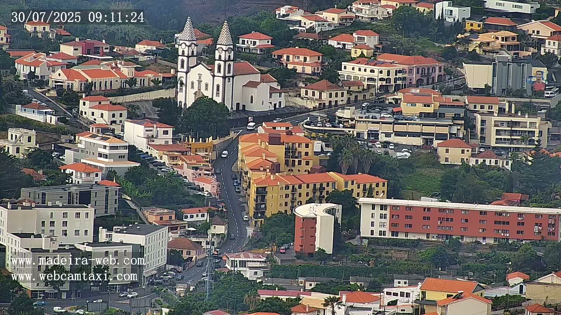 An aerial view of a town shows a white church with tall spires among numerous buildings, mostly low-rise with red tile roofs, on a slightly overcast day.