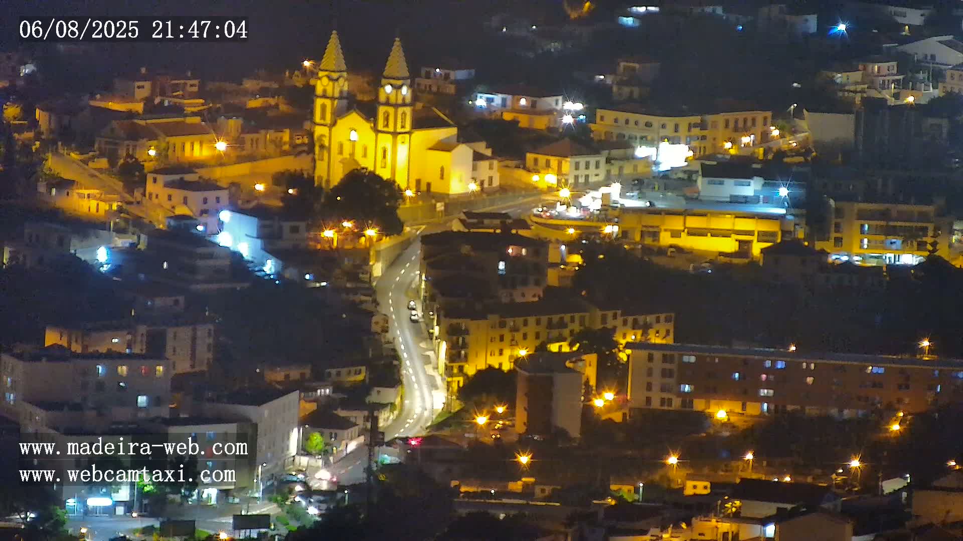 A nighttime aerial view of a town, illuminated by streetlights, featuring a prominent church and winding roads.
