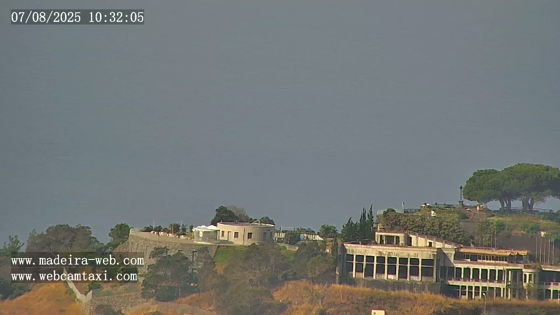 A hazy, overcast sky overlooks a hillside with several buildings, including a large, long structure, and some trees.