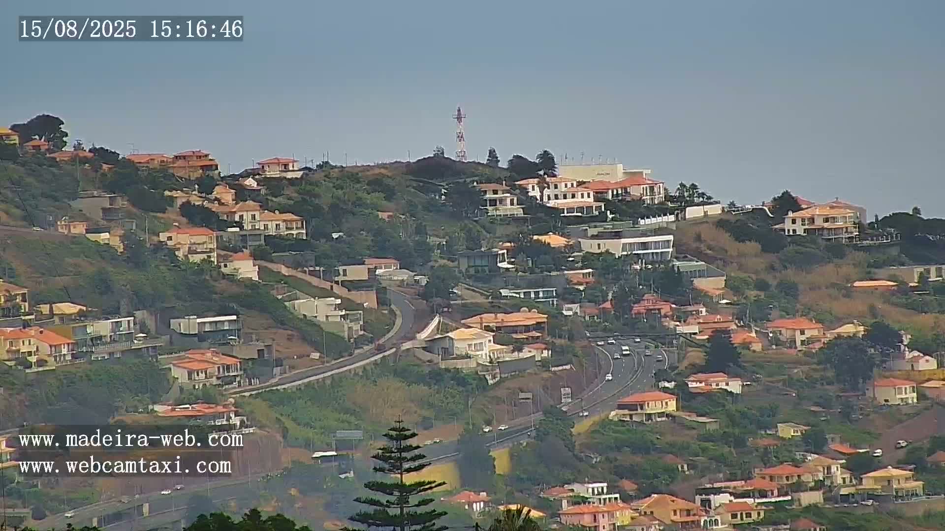 A hazy, overcast sky overlooks a hillside with several buildings, including a large, long structure, and some trees.