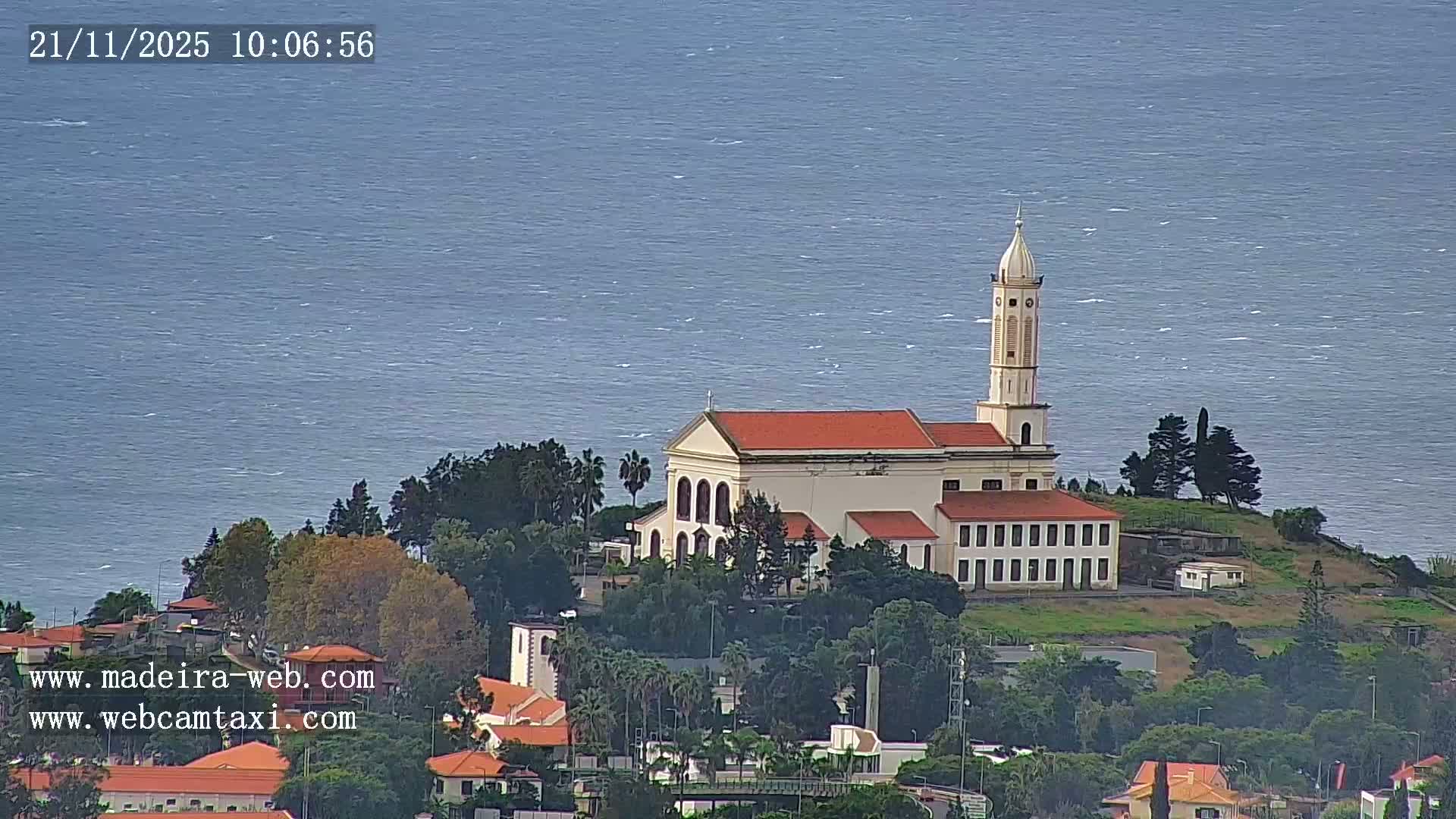 A large, light-colored church with a red roof and tall, domed steeple stands prominently on a lush green hillside, overlooking a choppy blue sea under what appears to be windy and overcast conditions.