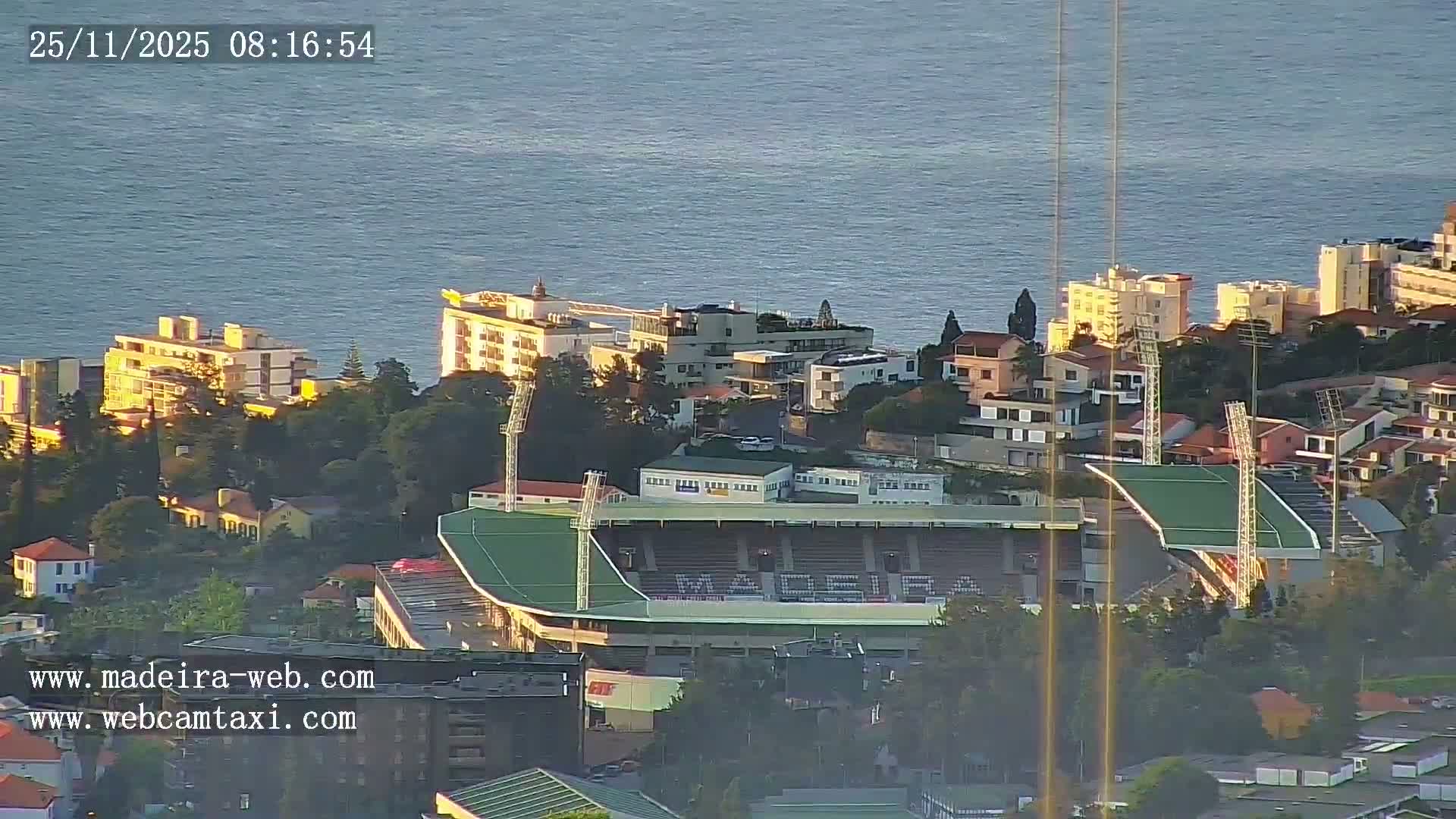 An aerial view reveals a coastal city with a prominent stadium nestled among buildings on sunlit hillsides, overlooking a calm blue sea under clear morning skies.