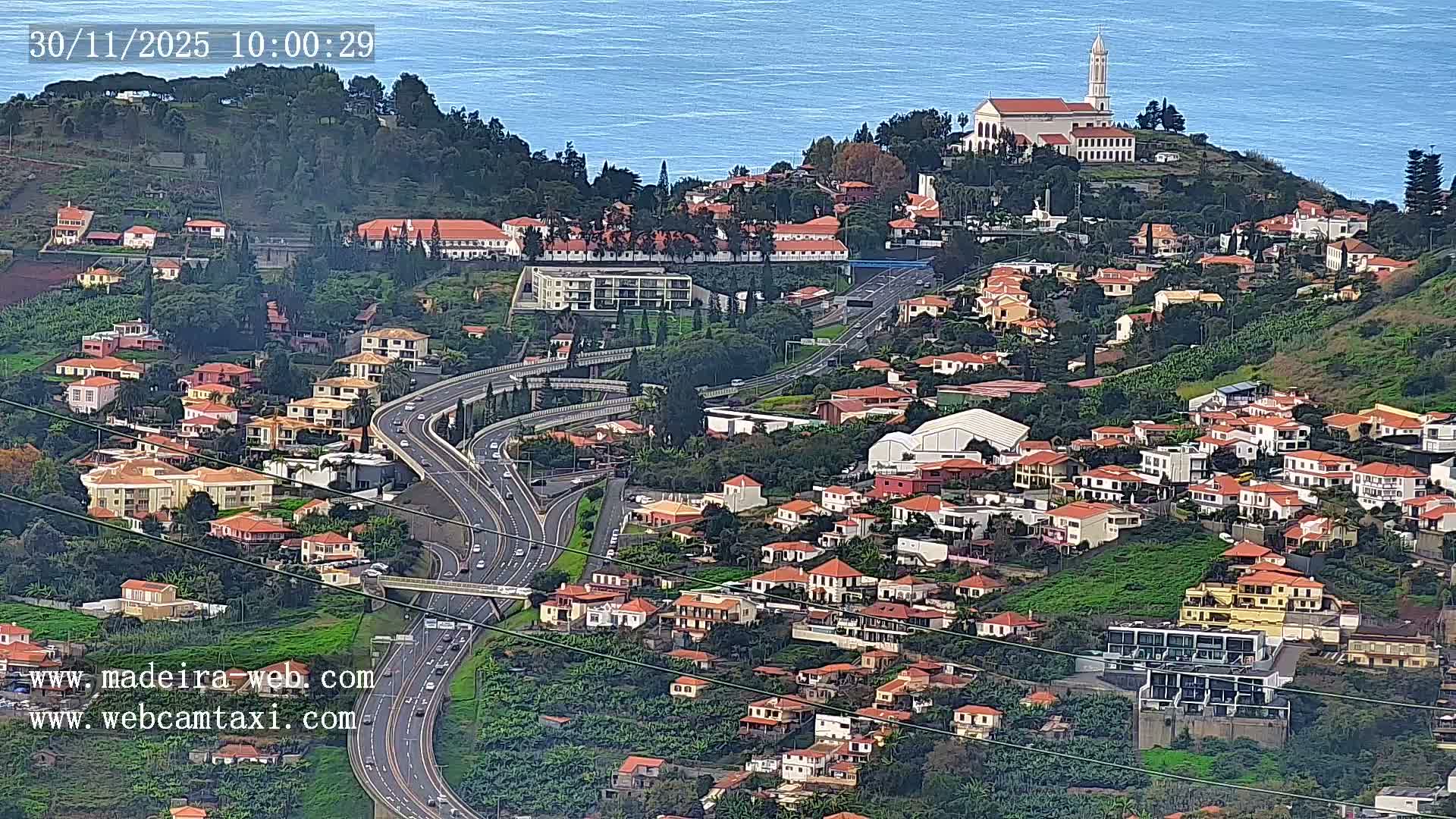 An aerial view reveals a town sprawling across lush green hillsides with numerous red-roofed buildings, a prominent church steeple, and winding highways, all set against a calm blue ocean under clear skies.