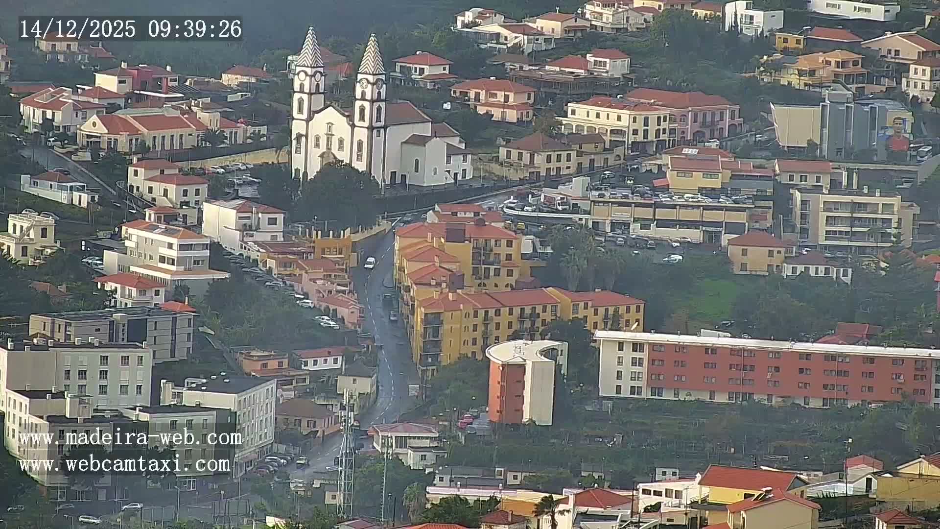 An aerial view reveals a town sprawling across lush green hillsides with numerous red-roofed buildings, a prominent church steeple, and winding highways, all set against a calm blue ocean under clear skies.