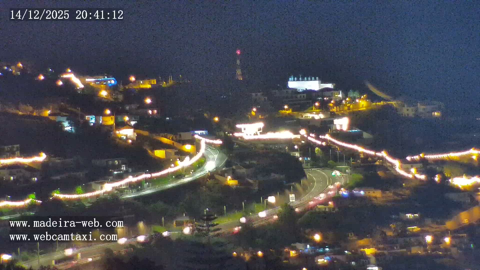 An aerial view reveals a town sprawling across lush green hillsides with numerous red-roofed buildings, a prominent church steeple, and winding highways, all set against a calm blue ocean under clear skies.
