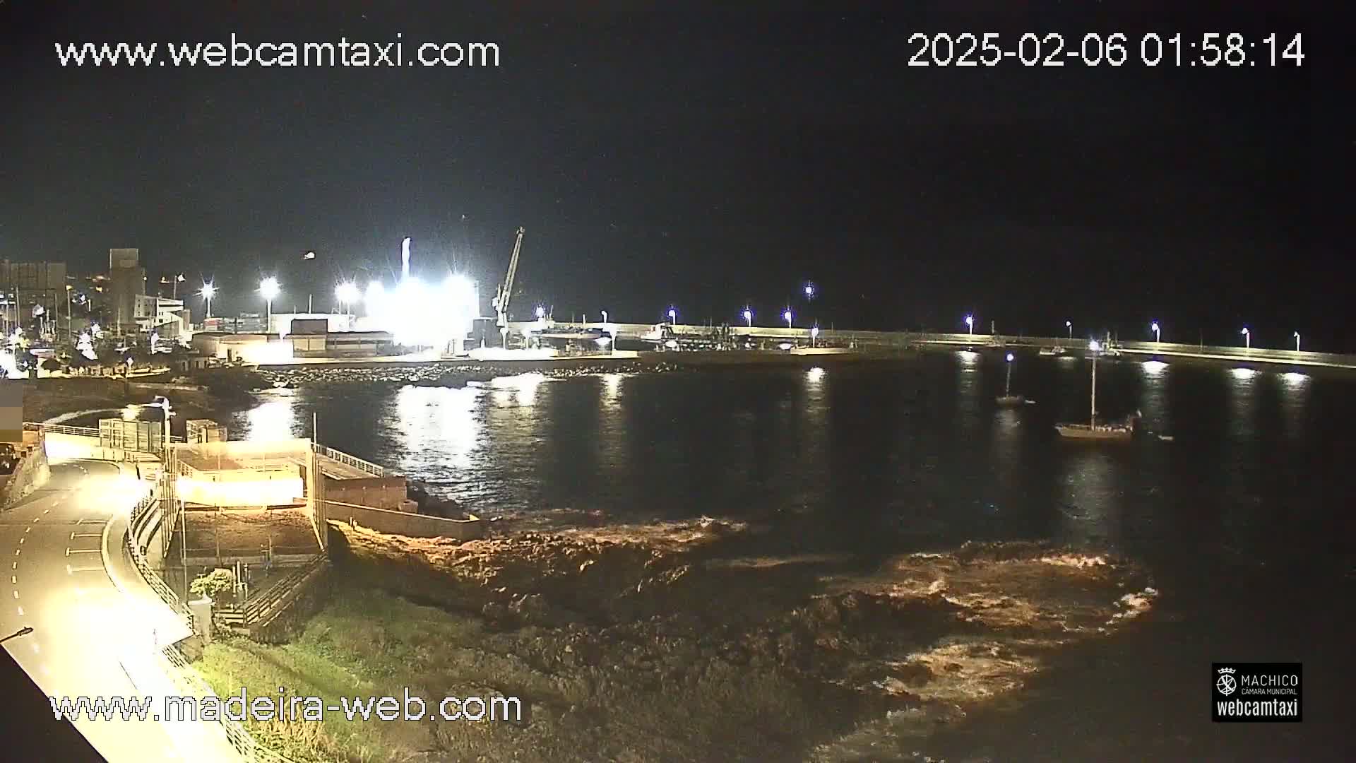 A nighttime view of a harbor with several boats, illuminated by artificial lights, and mild wave action near the shore.