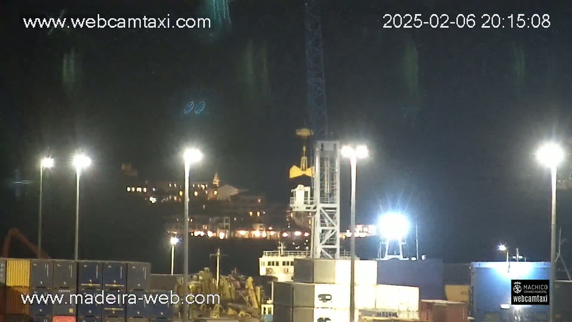 A nighttime view of a harbor with brightly lit cargo containers, a ship, and a crane under a dark, possibly cloudy sky.