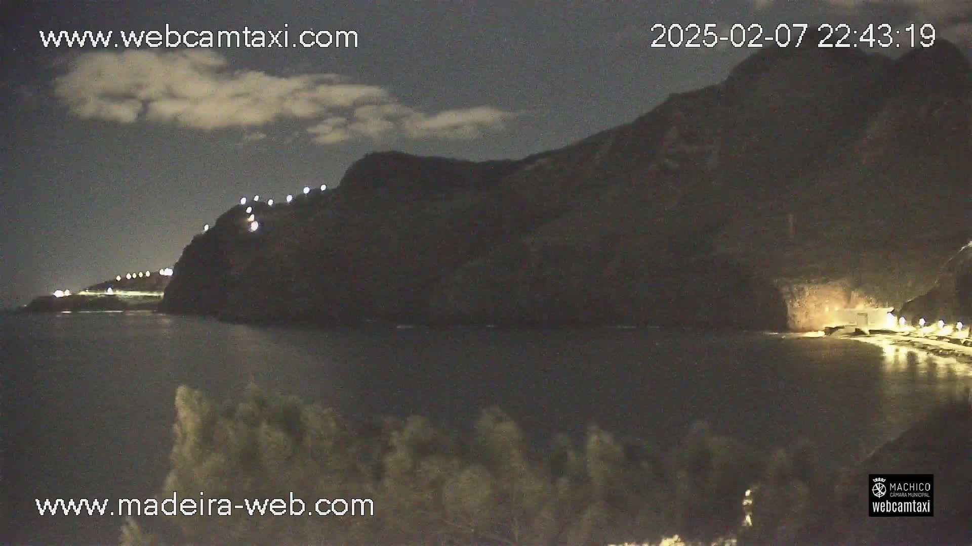 A nighttime coastal scene shows a dark, rocky cliff with lights along its edge, overlooking a calm body of water under a partly cloudy sky.