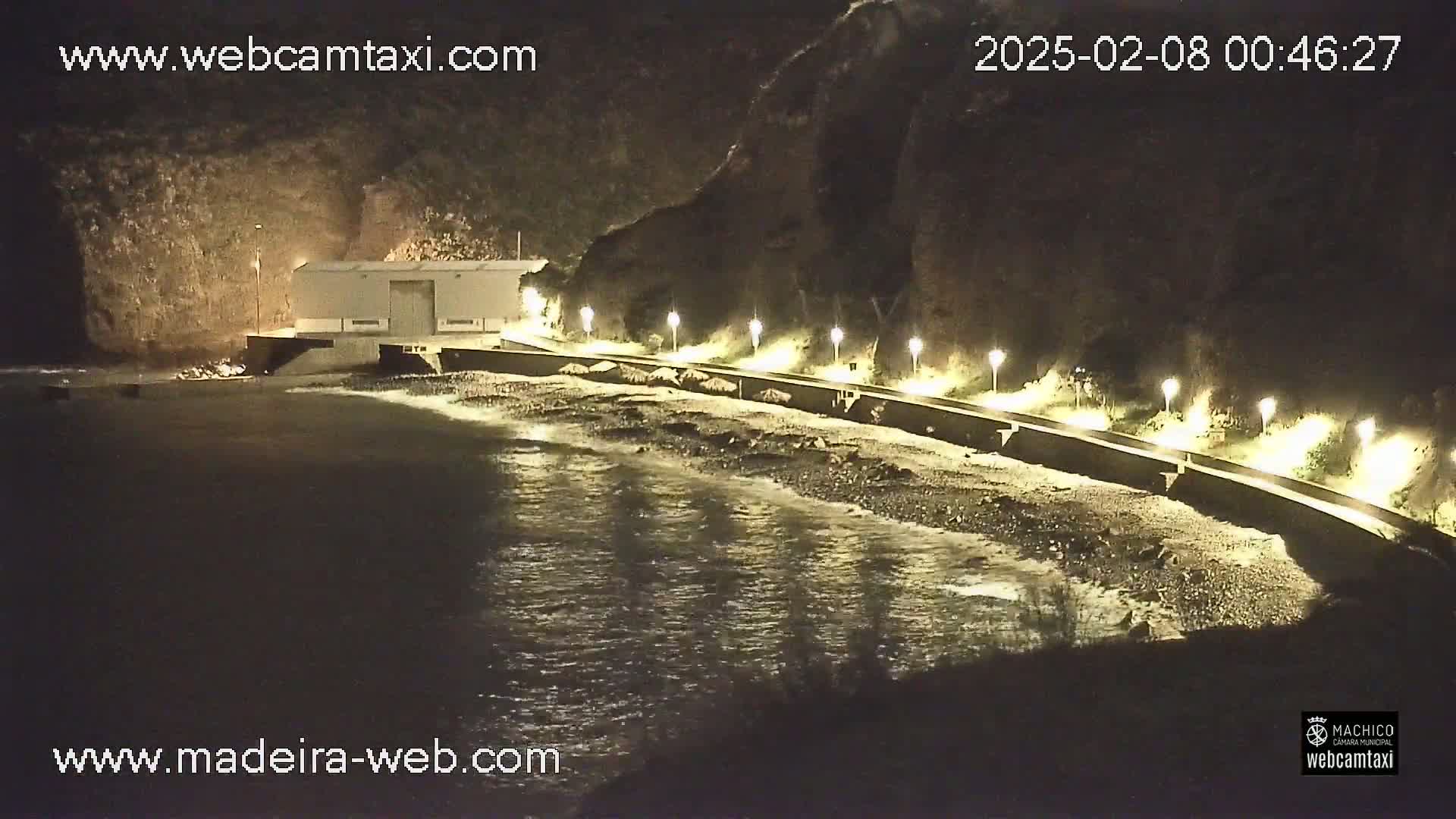 A nighttime coastal scene shows a building near a dark, rocky shore with gentle waves lapping at a pebble beach, illuminated by several lights along a walkway.