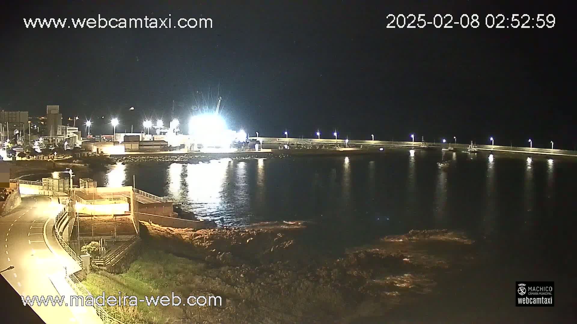 A nighttime view of a harbor with calm water, lit by artificial lights, showing a few boats and a structure on the waterfront.