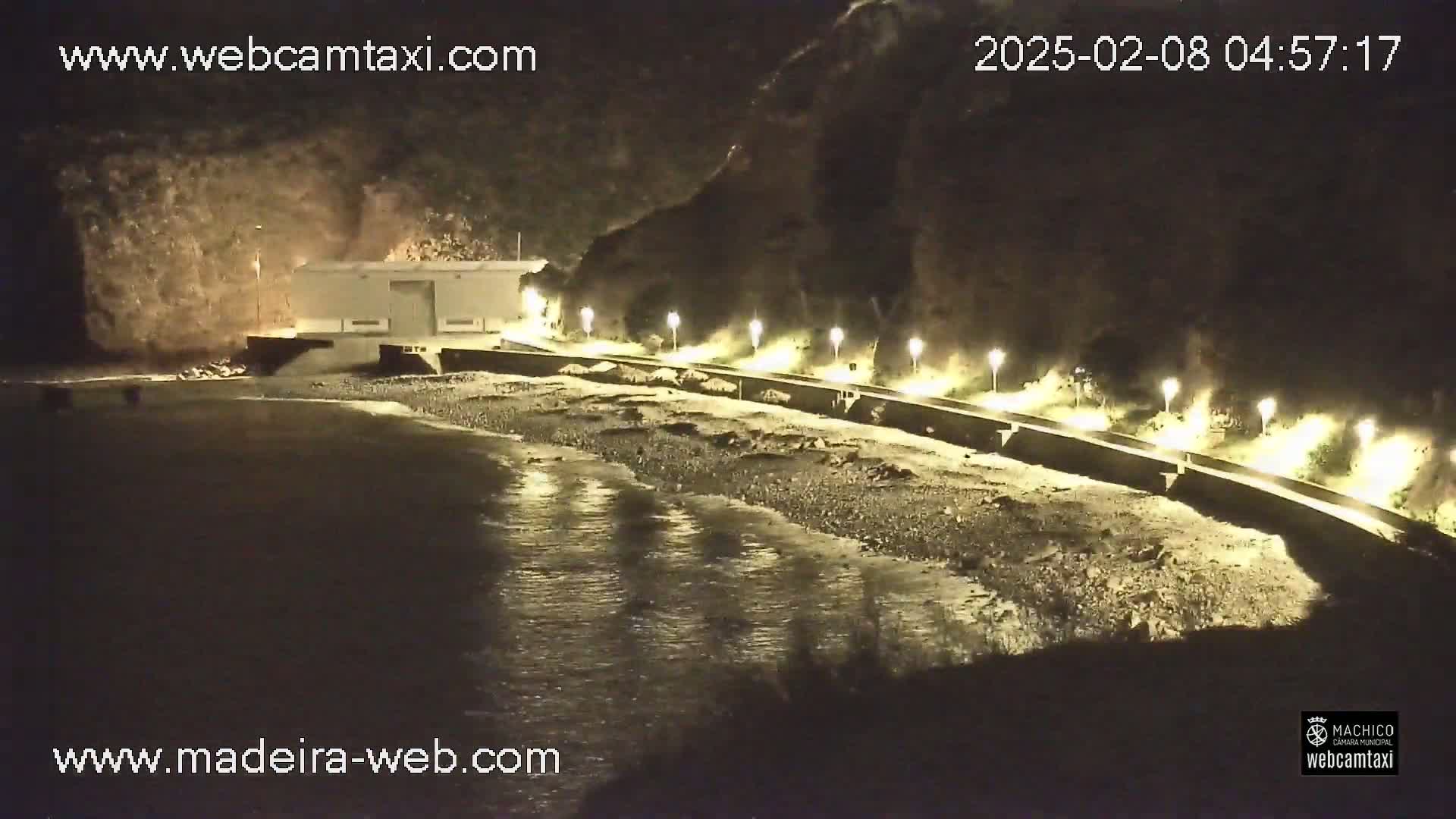 A nighttime, low-angle view shows a dark-colored body of water lapping against a rocky shore, with a lighted walkway and a building visible in the background.