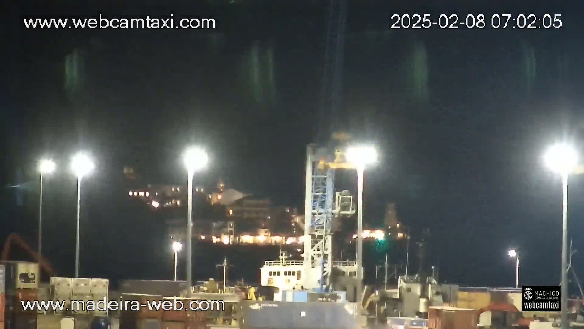 A nighttime view of a port with a large crane, a ship, and buildings illuminated in the background under a dark sky.