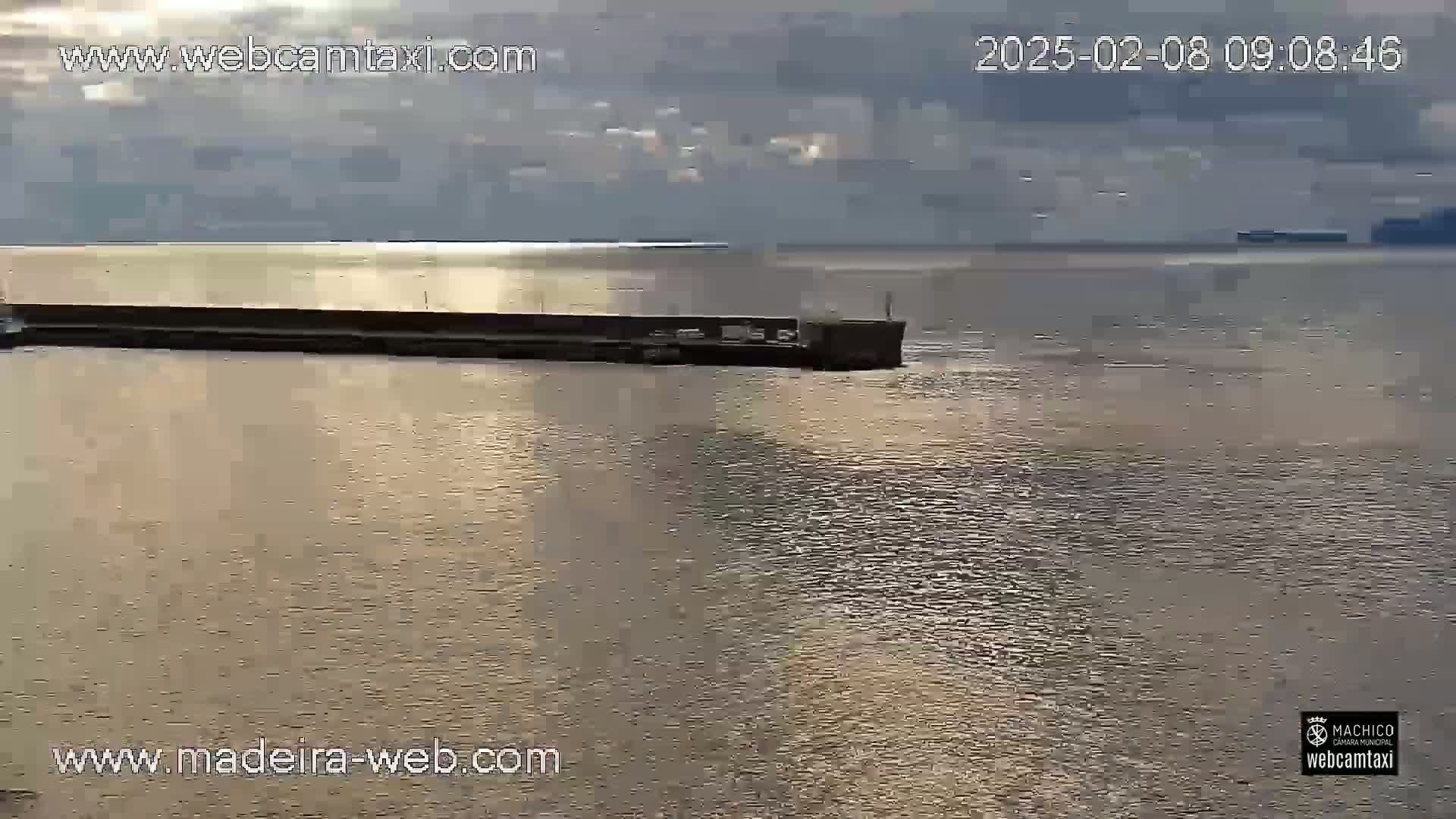 A long, low structure extends into a calm body of water under a mostly cloudy sky.