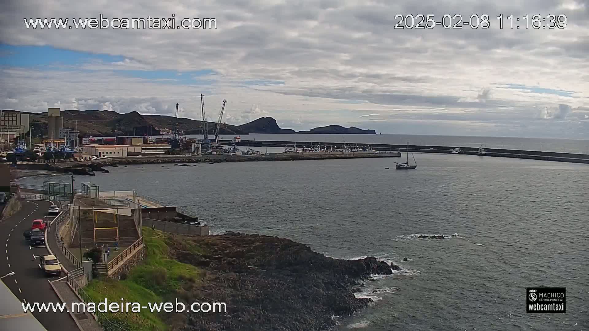 A harbor with several boats and industrial buildings is seen under a mostly cloudy sky.