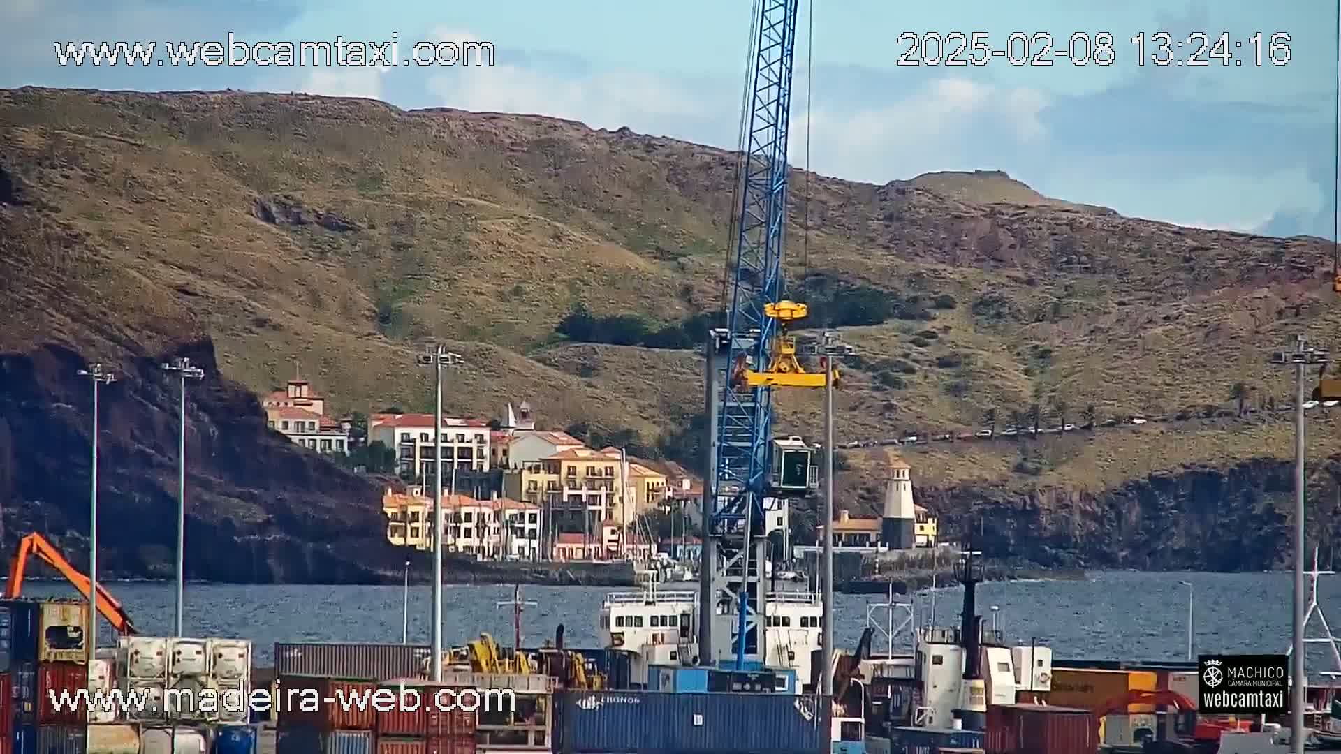 A harbor scene under partly cloudy skies shows a large blue crane in the foreground, cargo containers, a small town nestled against a rocky hillside, and a lighthouse on a small headland.