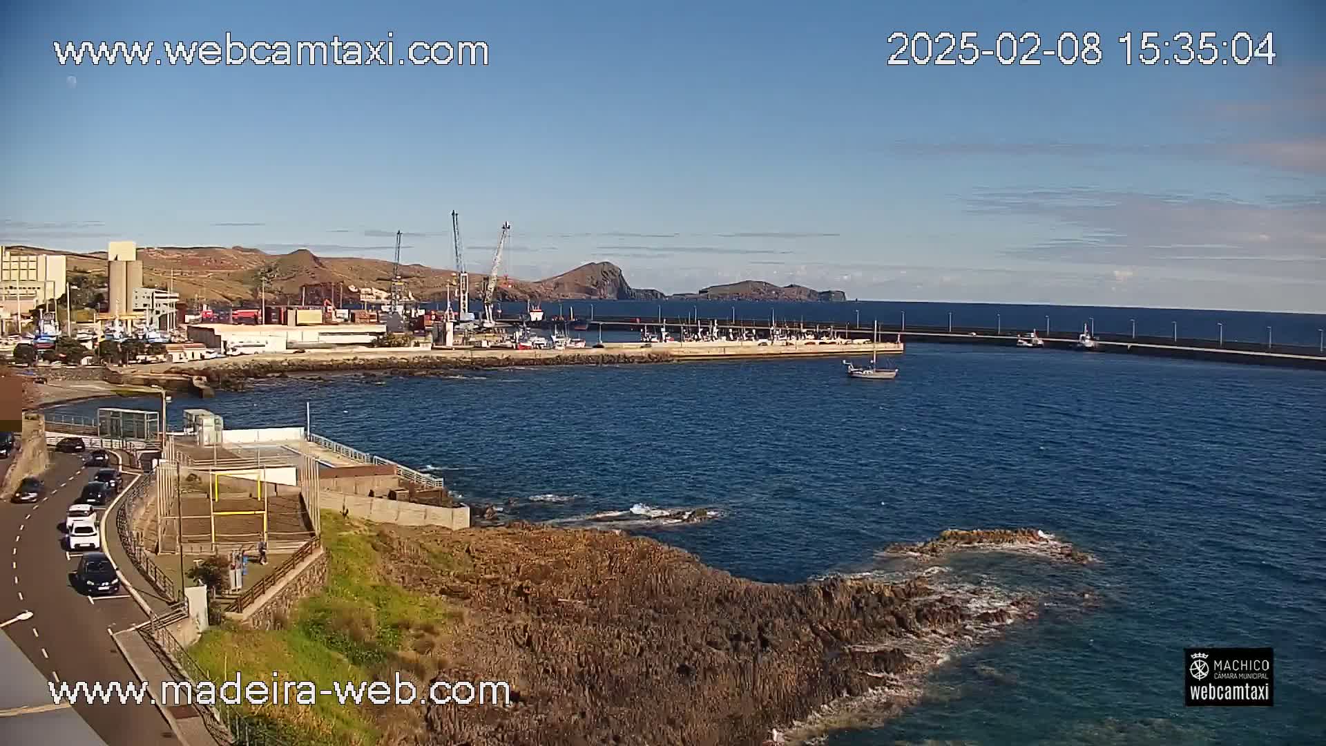 A harbor filled with boats is seen from a high vantage point on a sunny day, with a rocky coastline in the foreground and hills in the background.