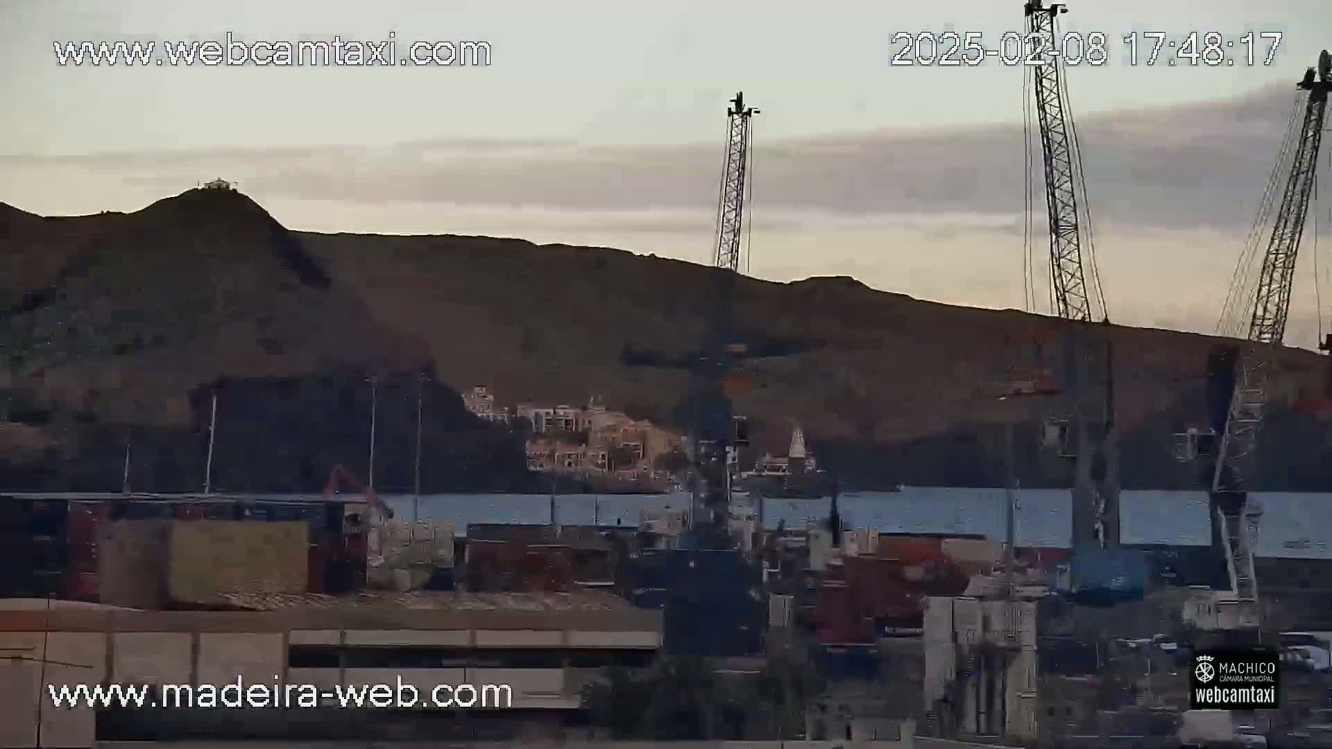 A harbor scene at dusk shows several cranes and cargo containers near a body of water with a large, dark hill in the background.