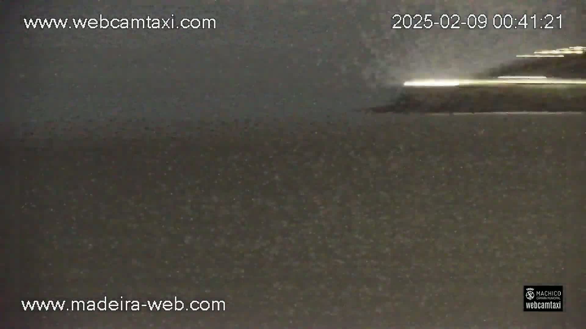 A dark, possibly nighttime, outdoor scene shows a blurry, light-colored object moving across a dark surface under what appears to be light rain or snow.