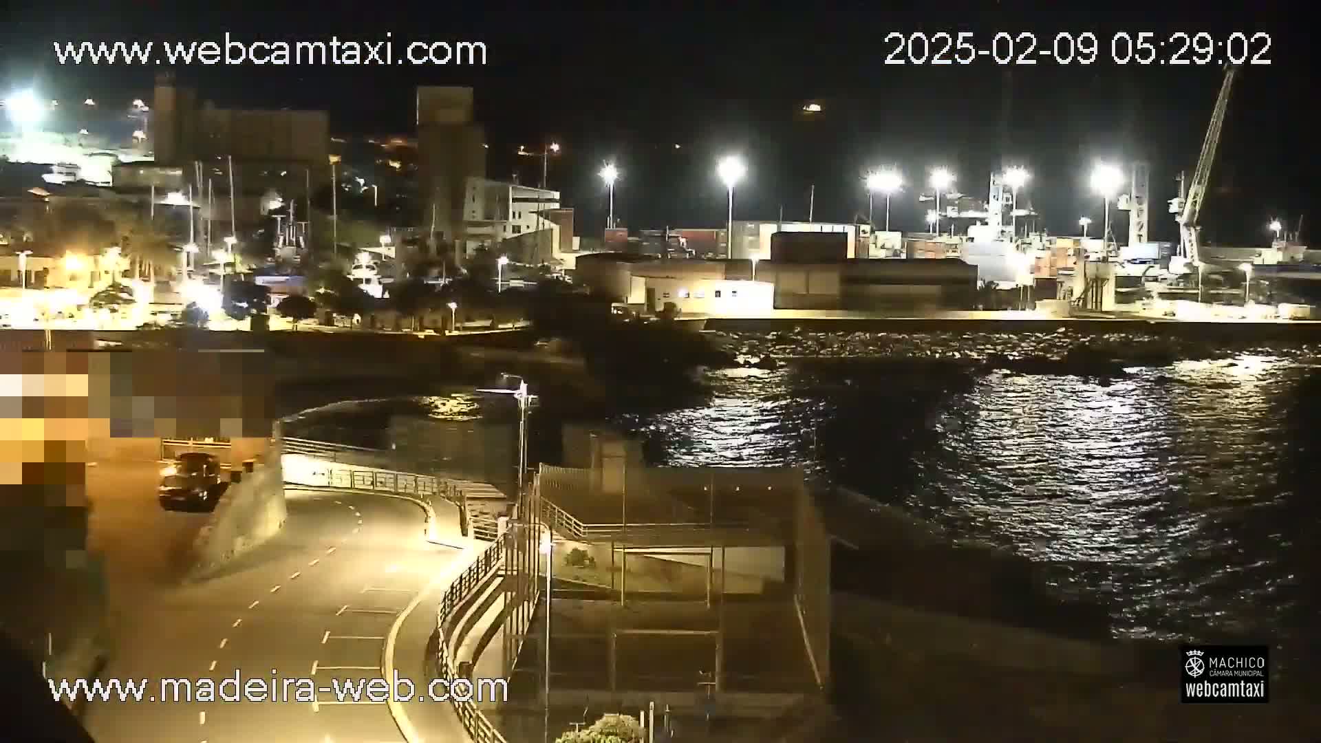 A nighttime view of a harbor with calm water, showing a roadway curving along the waterfront next to buildings and industrial areas.