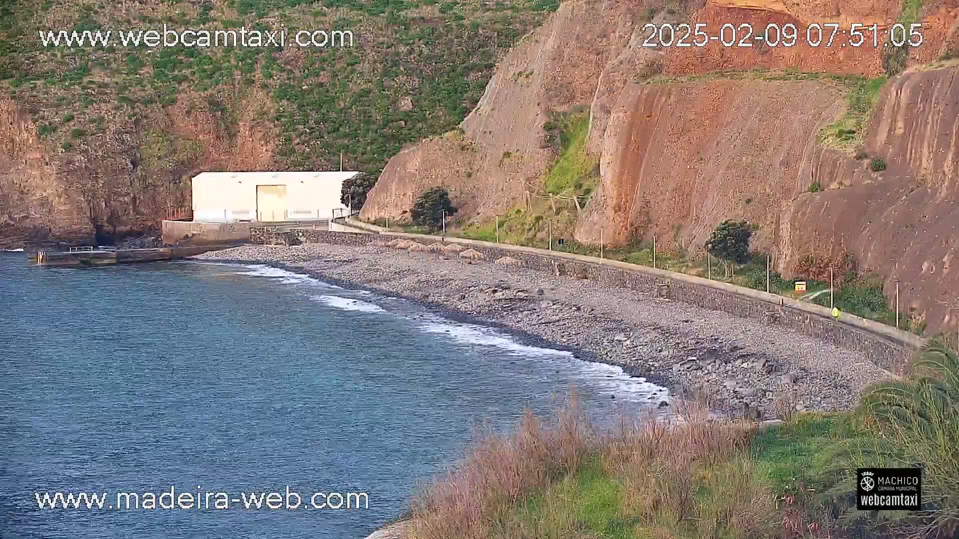 A rocky beach with a white building and a paved walkway next to a steep, reddish-brown cliff faces a calm ocean.