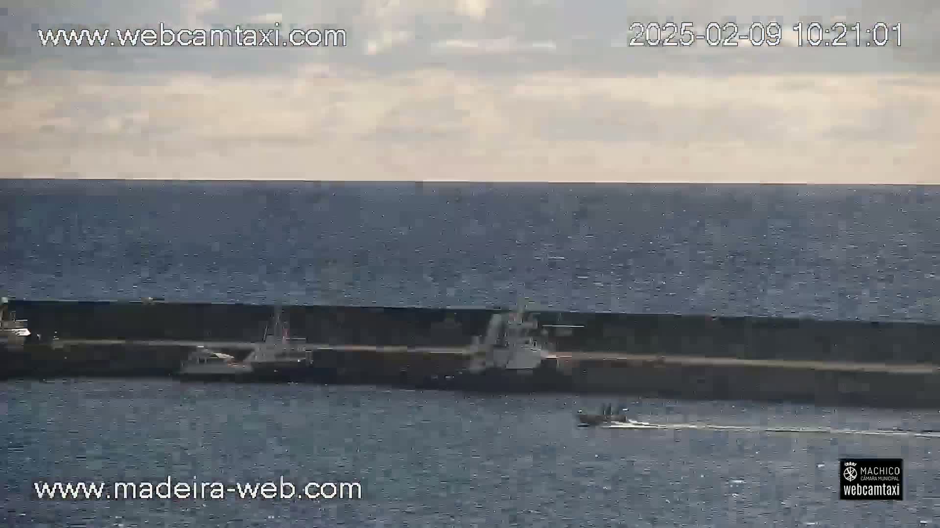 Several small vessels are moored at a dock along a calm, dark-blue ocean under a mostly cloudy sky.