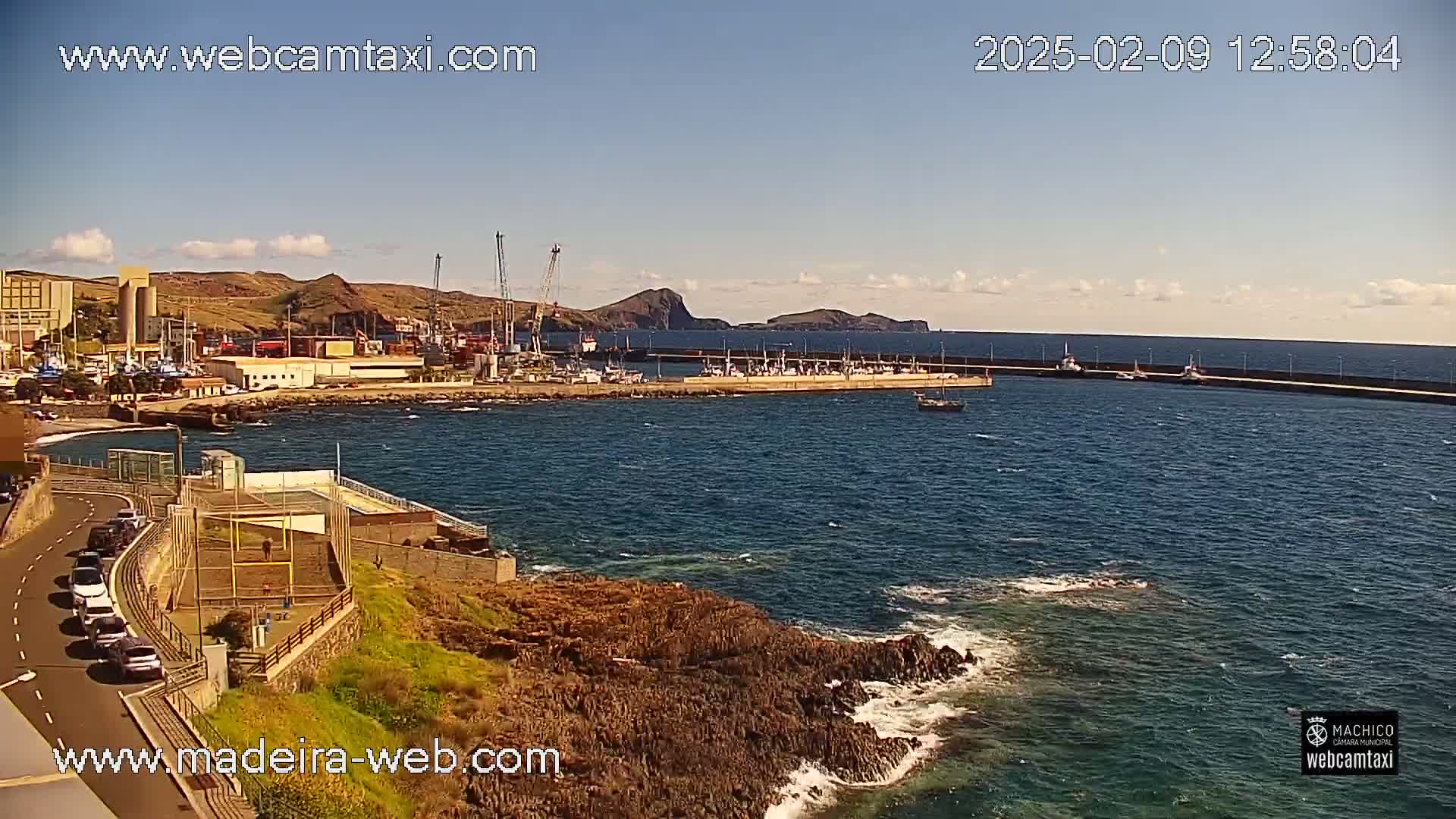 A harbor filled with boats is seen from a high vantage point on a sunny day with some clouds, overlooking a rocky coastline with light wave action.