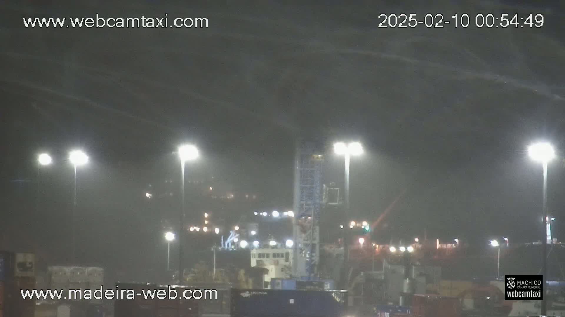A nighttime view of a harbor with several ships and lights, under a hazy, possibly rainy or foggy sky.
