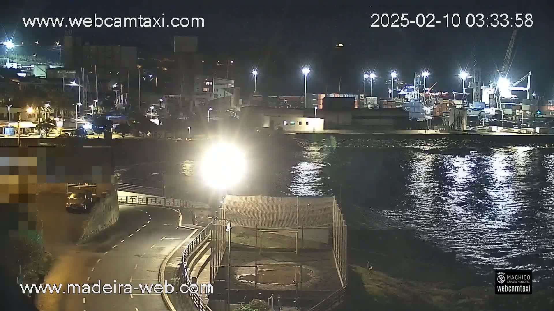 A nighttime view of a harbor with boats and a cargo area, next to a road and a fenced-in area, under bright lights, with calm water.