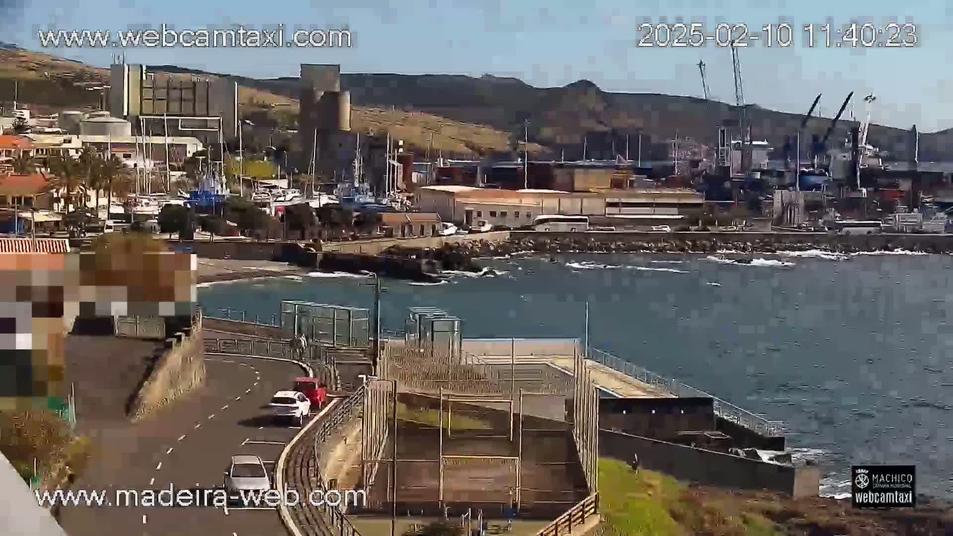 A sunny day overlooking a harbor with boats, buildings, and a roadway, featuring a few cars traveling along the road.