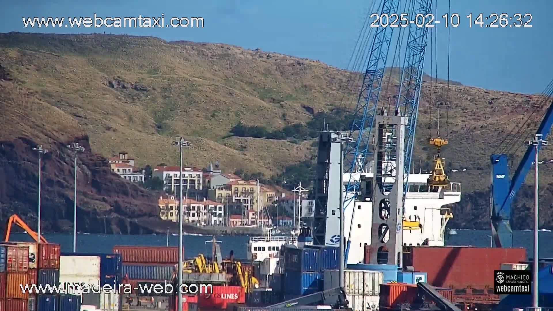 A cargo ship is being loaded at a port under a clear blue sky, with a hilly landscape and buildings in the background.