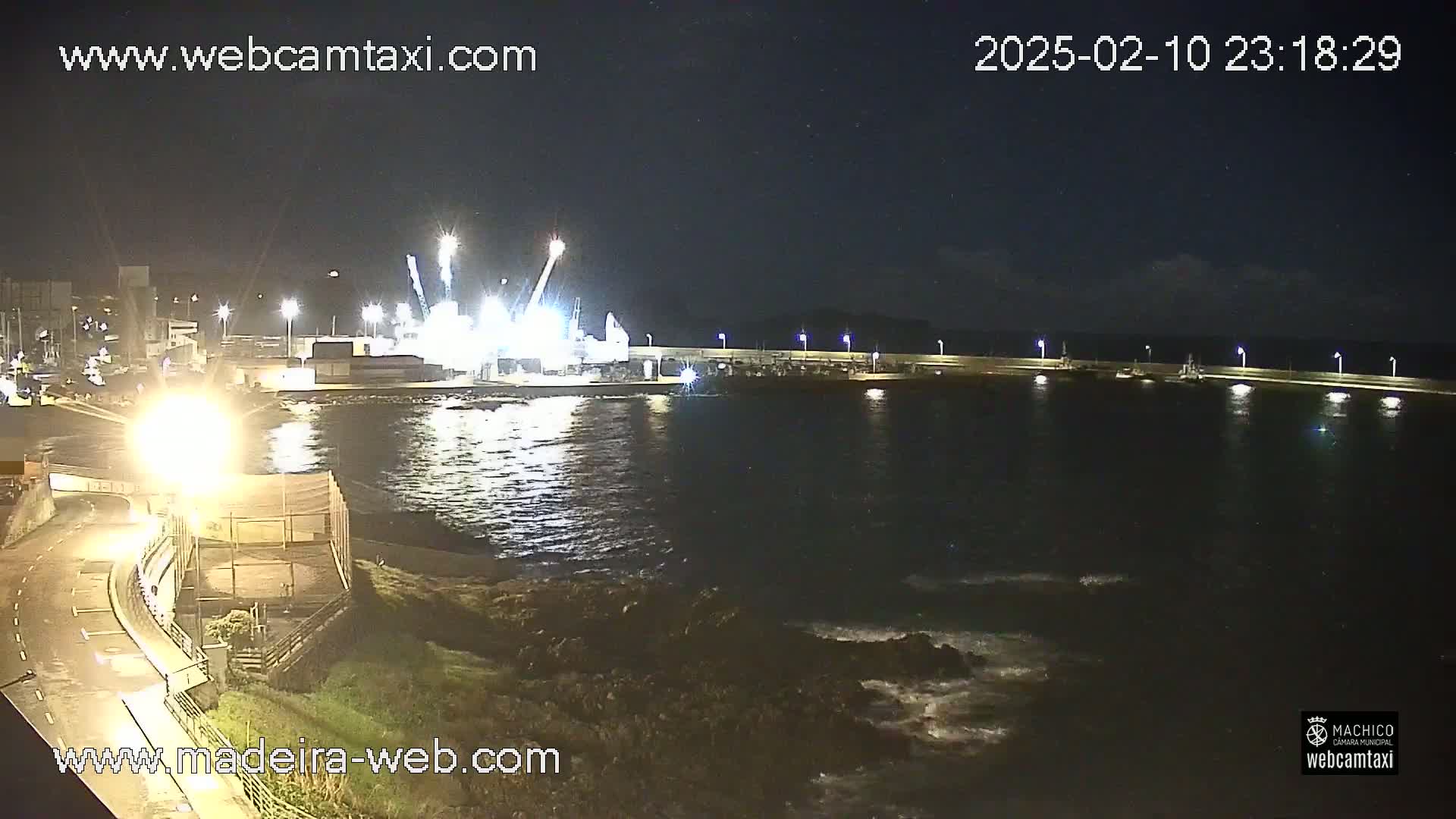 A nighttime view of a harbor with brightly lit cranes and other industrial equipment on a pier, with slightly choppy water lapping against the rocky shore.