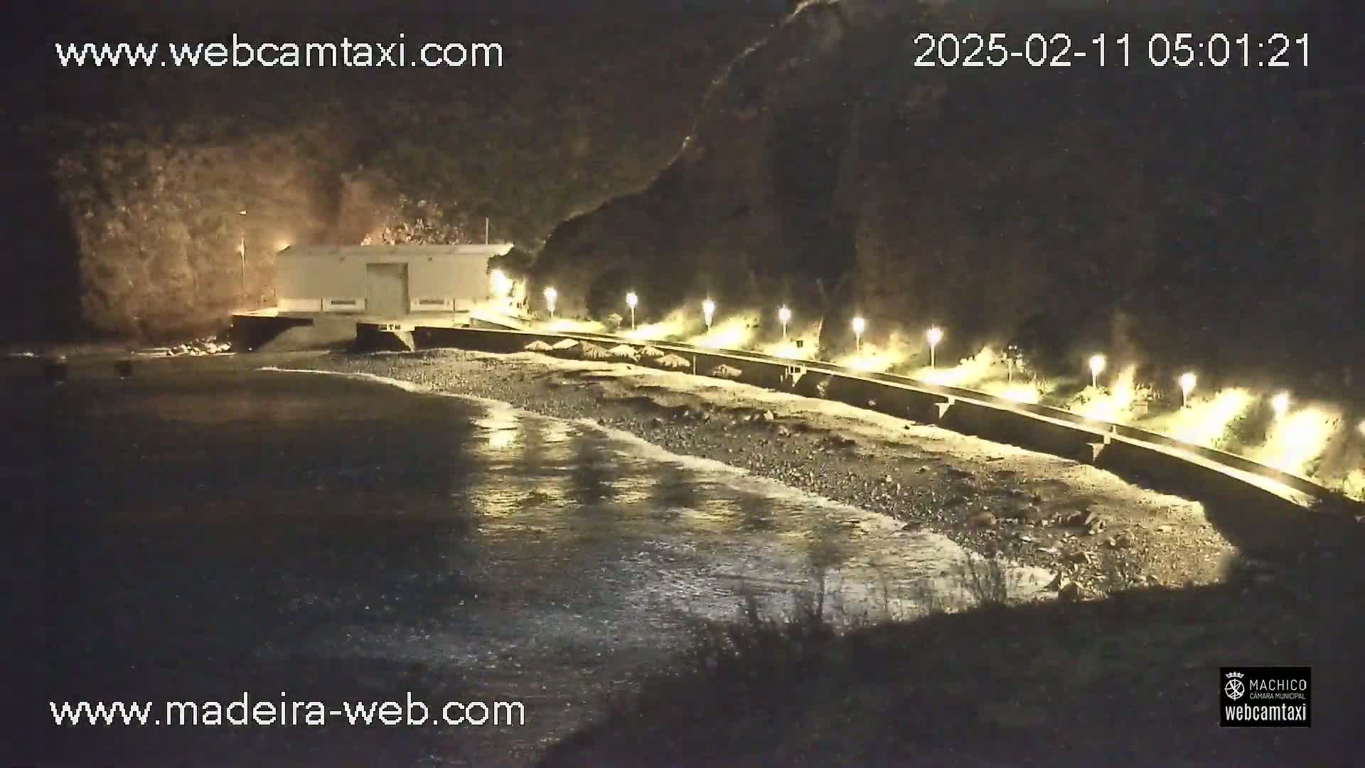 A nighttime view of a rocky beach with gentle waves lapping the shore, illuminated by lights along a seawall that runs beside a building nestled against a dark cliff face.