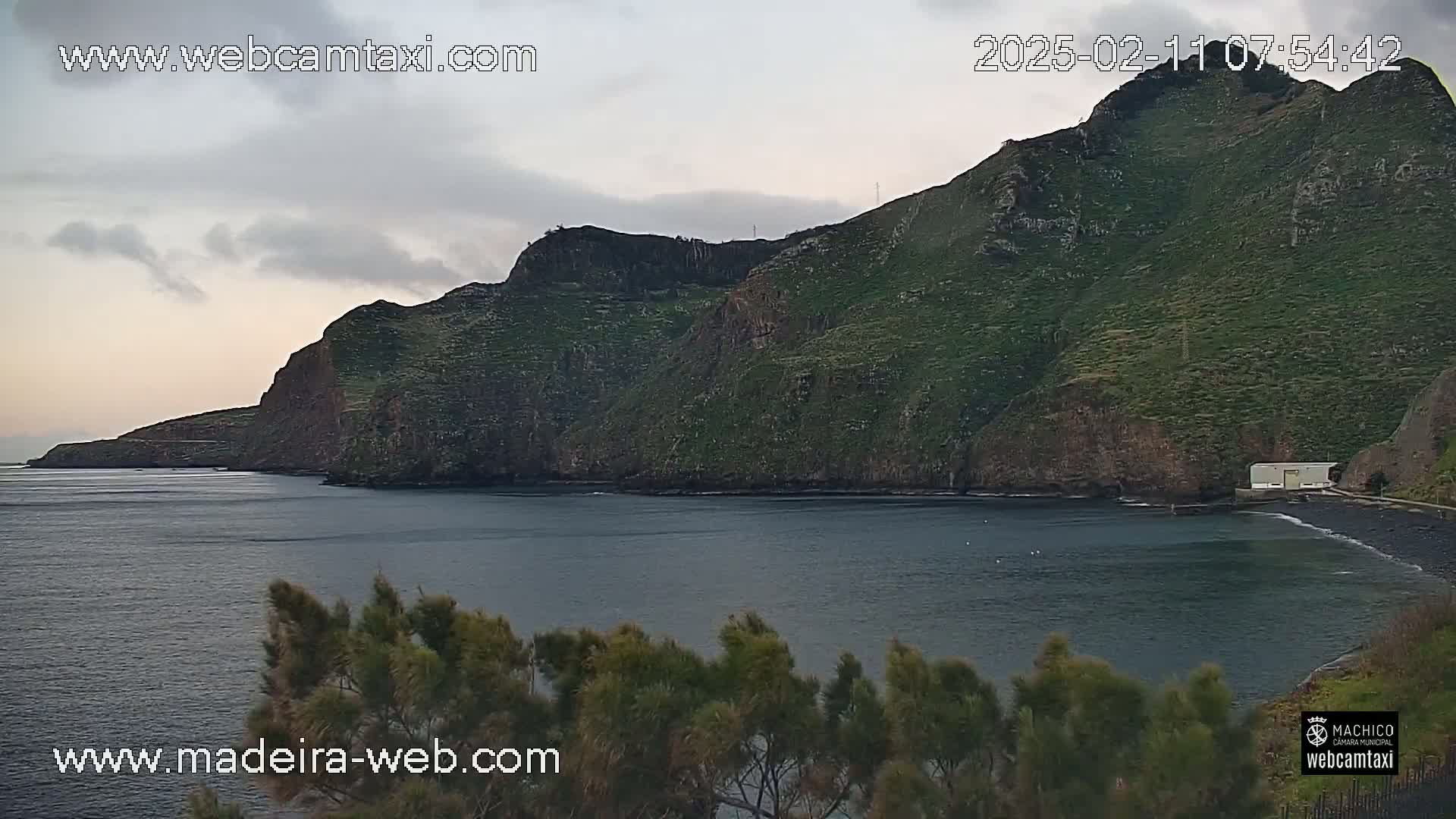 A dark-grey, overcast sky hangs over a calm body of water next to a steep, green-covered cliff.