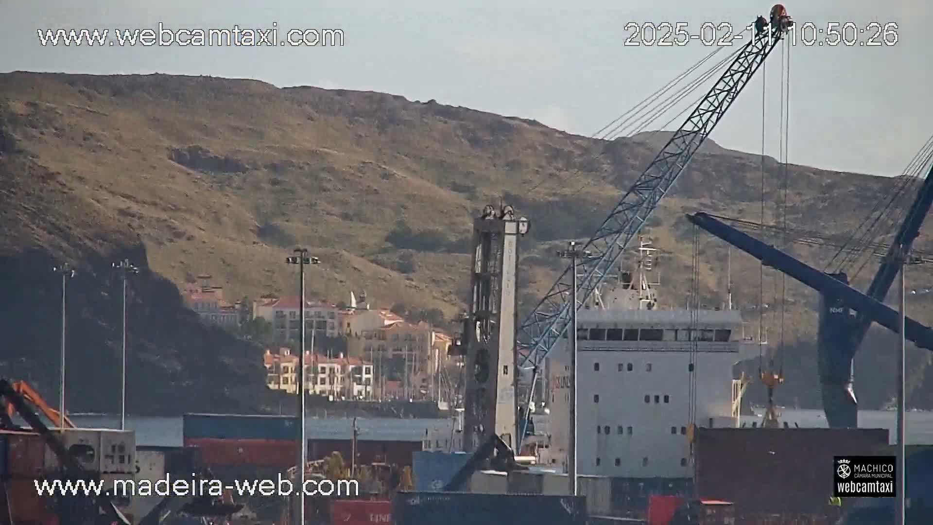 A large cargo ship is docked at a port under a mostly clear sky, with a hill in the background and several cranes visible.