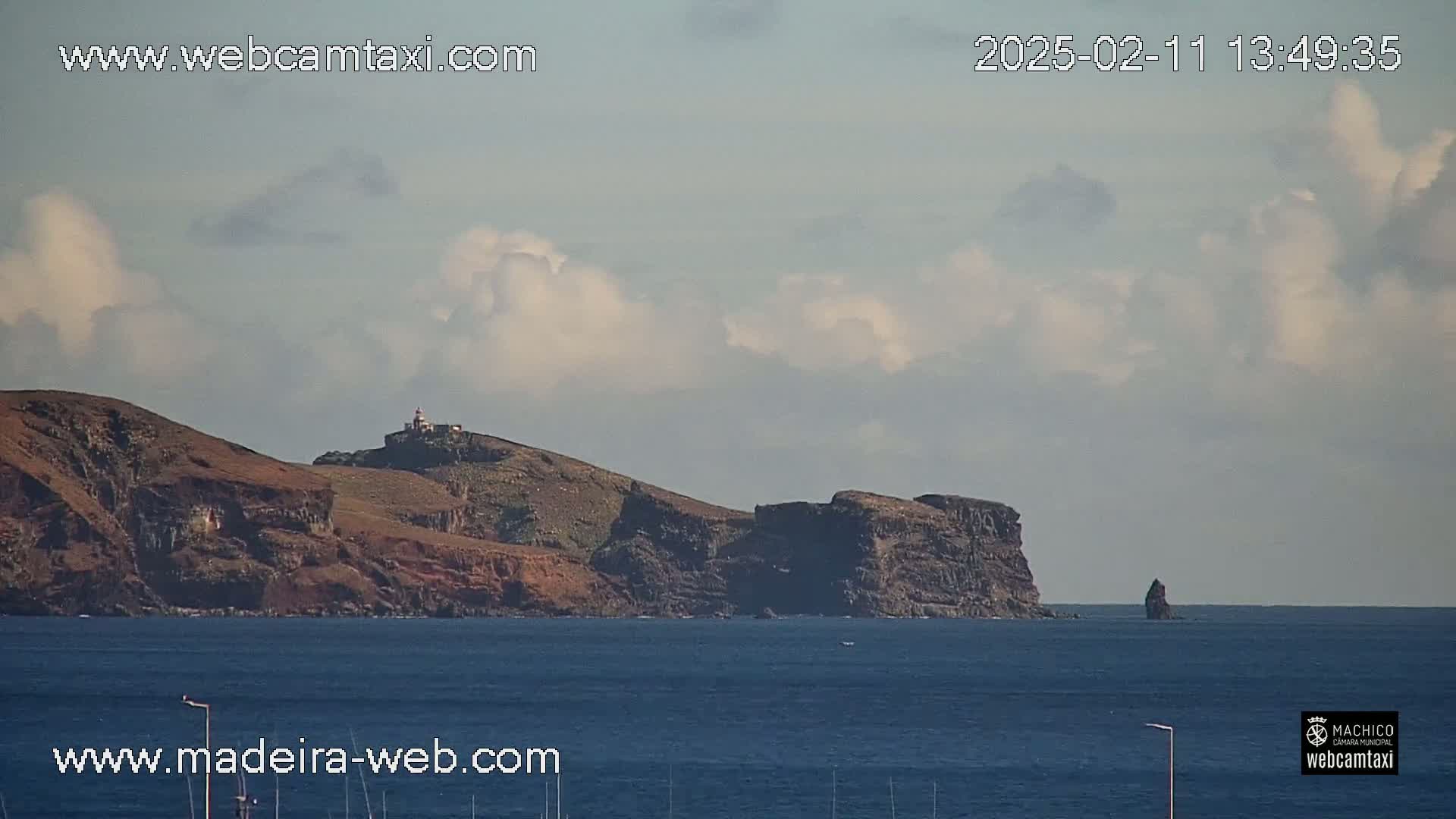 A rocky, hilly coastline with a lighthouse is visible under a partly cloudy sky over a dark blue ocean.