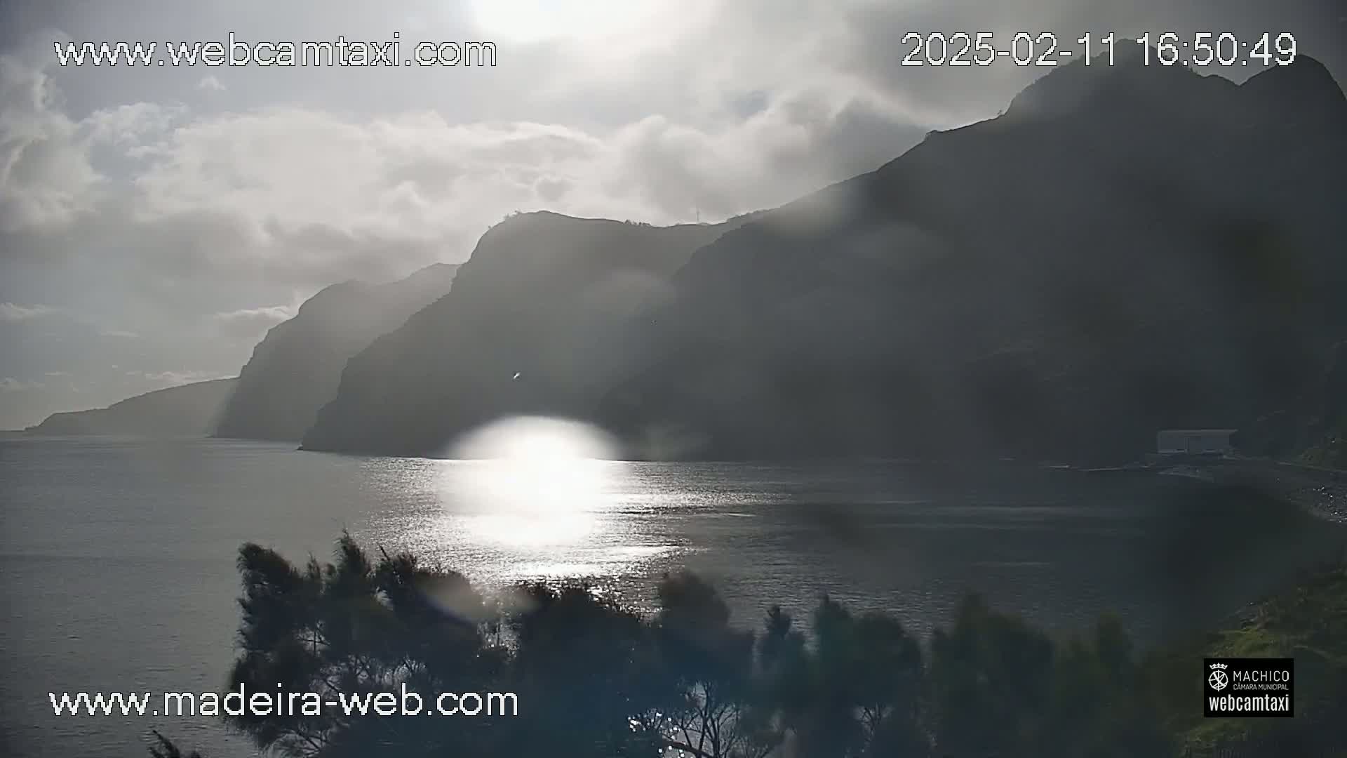 A partly cloudy day reveals a sunlit ocean reflecting off the dark, steep cliffs of a coastline.