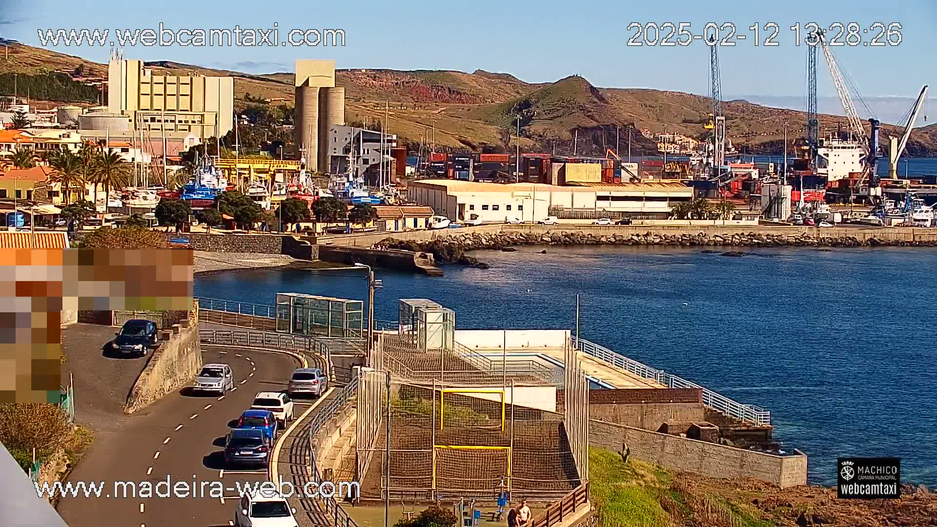 A harbor scene on a sunny day shows a coastal town with buildings, boats, and a small sports field next to the water, along with cars driving on a nearby road.