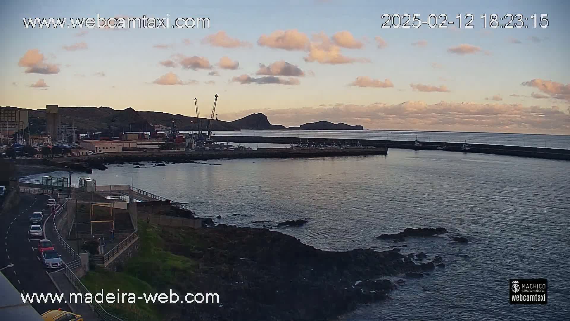 A harbor with calm water under a partly cloudy sunset sky, showing a town, port, and rocky coastline.