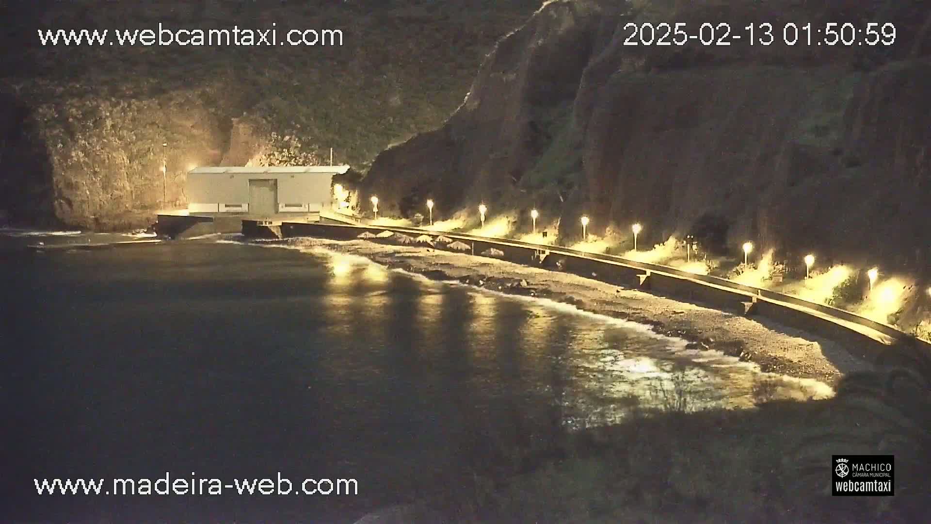 A nighttime view of a calm, dark-colored body of water next to a pebble beach, illuminated by lamps lining a seawall beneath dark cliffs.