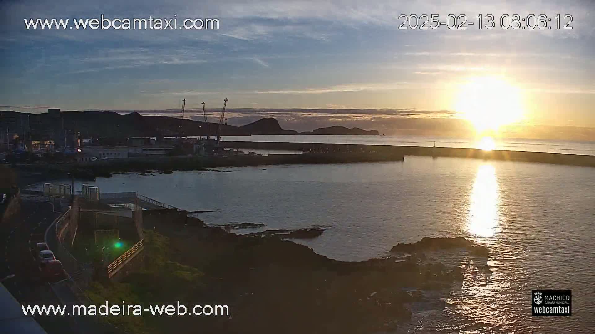 A sunrise over a calm harbor, with a town and hills in the background.