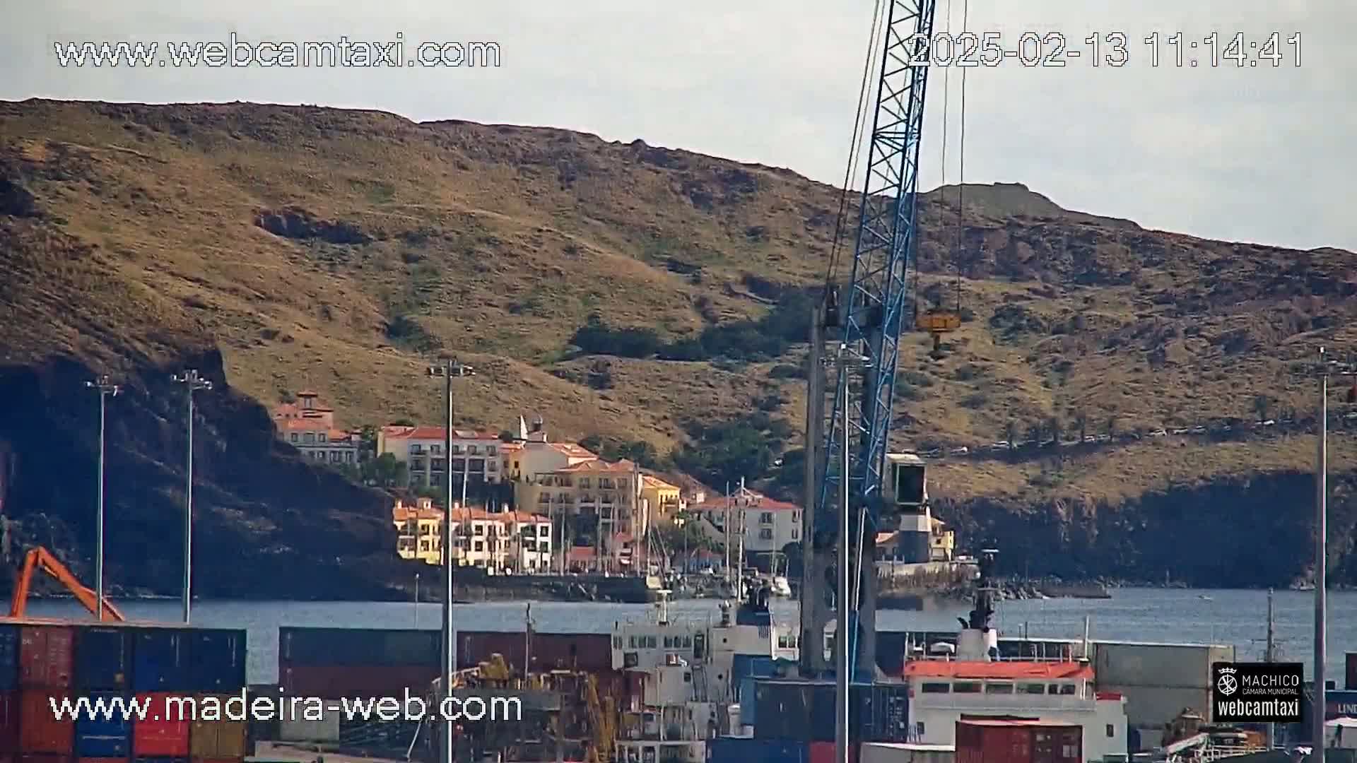A harbor scene with a large crane, cargo containers, buildings on a hillside, and calm water under a mostly clear sky.