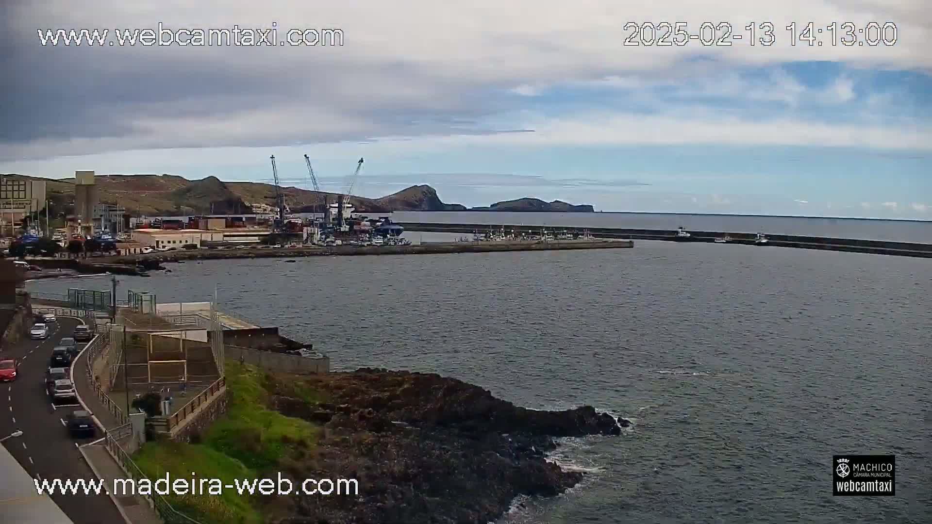 A partly cloudy day reveals a harbor with docked ships and cranes, a roadway with cars, and a rocky coastline meeting calm water.