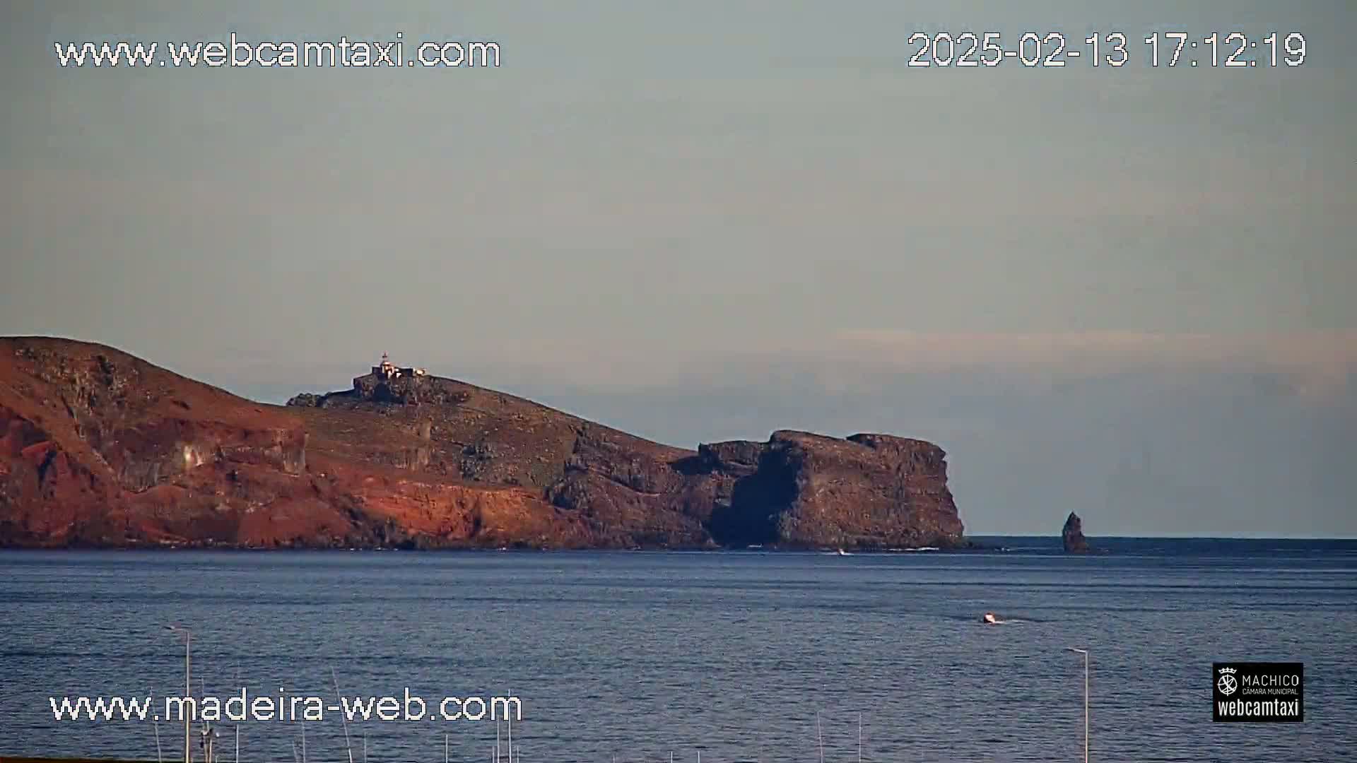 A distant rocky island with a lighthouse atop a hill is seen across a calm body of water under a mostly clear sky.