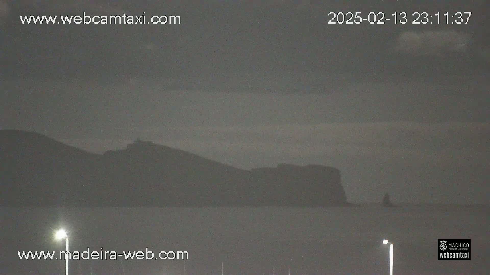 A dark, overcast nighttime scene shows a silhouetted mountain range across a body of calm water, with two lit lampposts in the foreground.