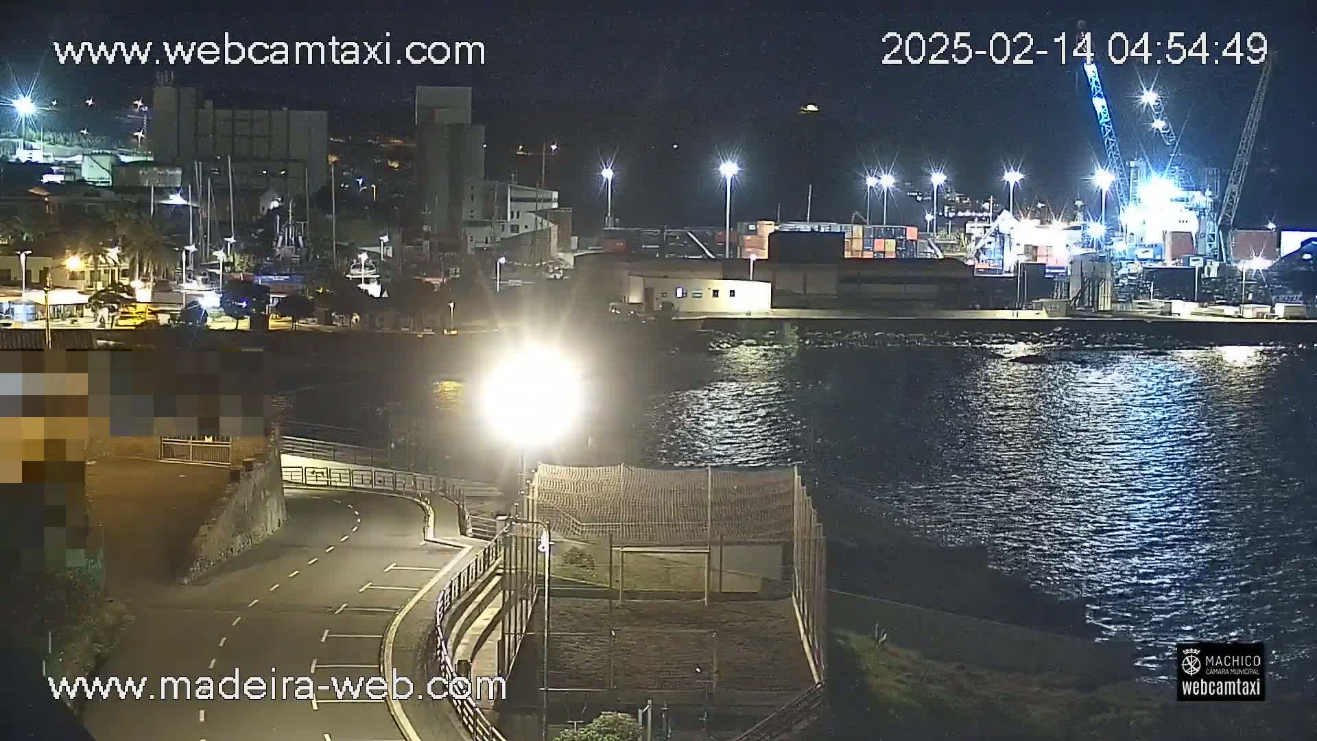 A nighttime view of a harbor with calm water, brightly lit docks, and several ships, under a clear sky.