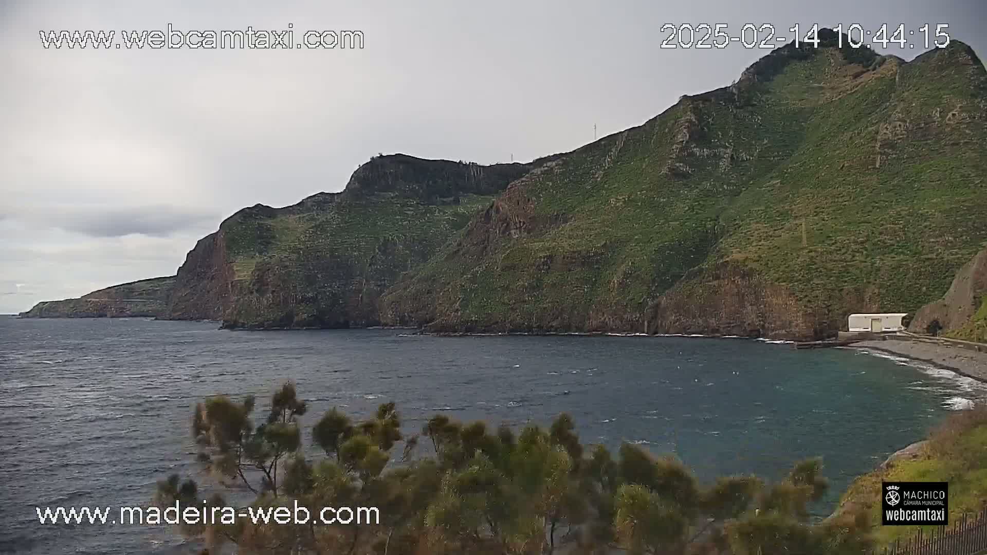 A dark-grey overcast sky hangs over a calm, dark-blue ocean partially enclosed by a steep, green-covered cliff face.