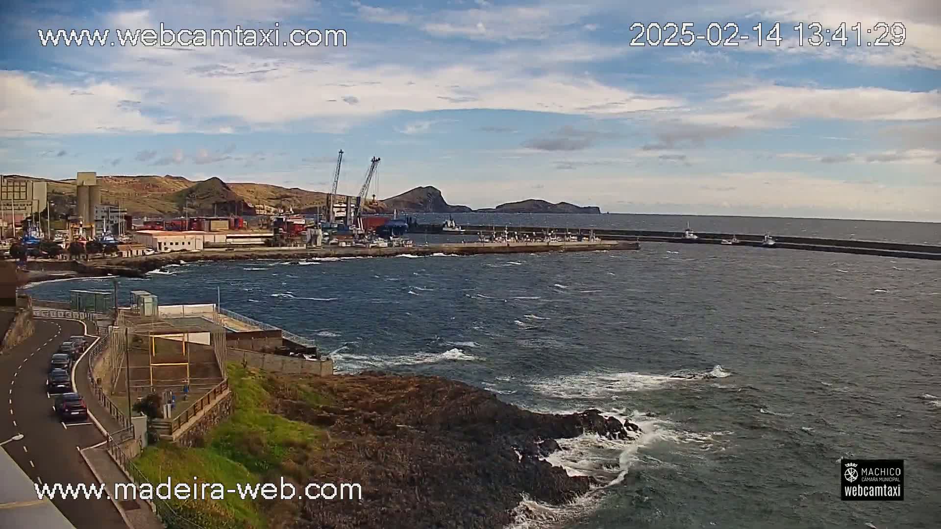 A harbor with several boats and cranes is visible from a rocky coastline with a slightly choppy sea under a partly cloudy sky.